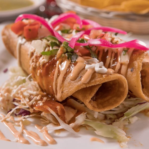 Two golden-brown taquitos topped with red onions, salsa, and creamy sauces on a bed of shredded lettuce.