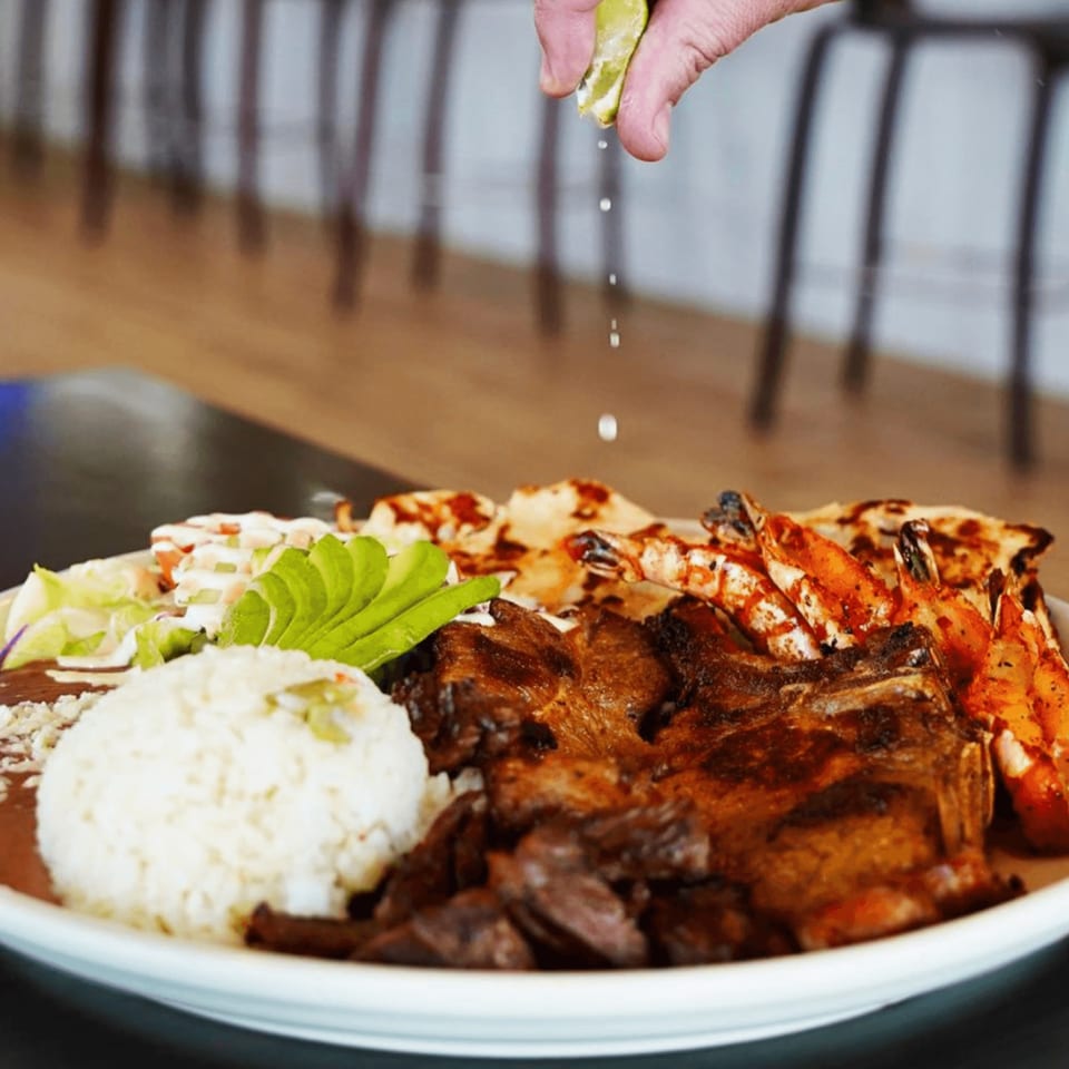 A hand squeezes lime over a Lempira Restaurant platter of grilled meat, shrimp, rice, beans, and salad.