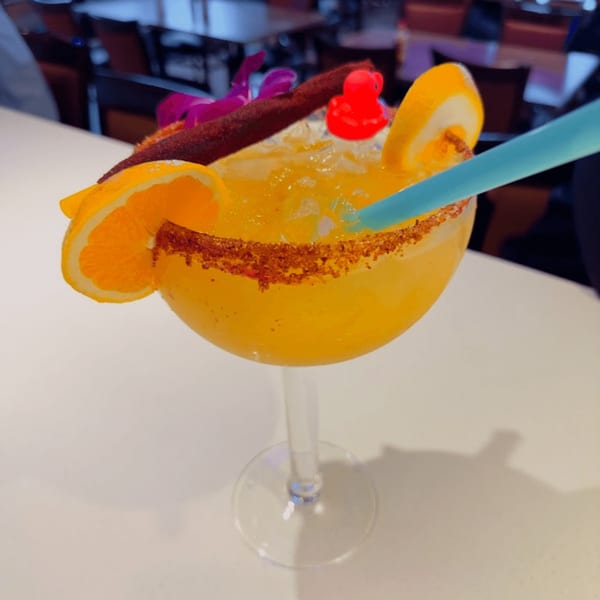 A vibrant orange margarita with a chili-lime rim, orange slices, a red rubber duck, and a blue straw.