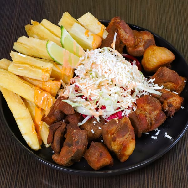 A black plate filled with fried yucca, crispy pork chunks, and a fresh cabbage salad.
