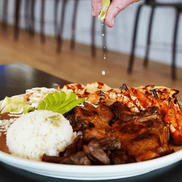 Hand squeezing lime over a Lempira Restaurant platter with grilled meat, shrimp, rice, and avocado.