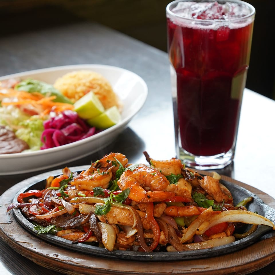 Sizzling fajitas with shrimp, chicken, onions, and peppers, alongside a salad and red drink.